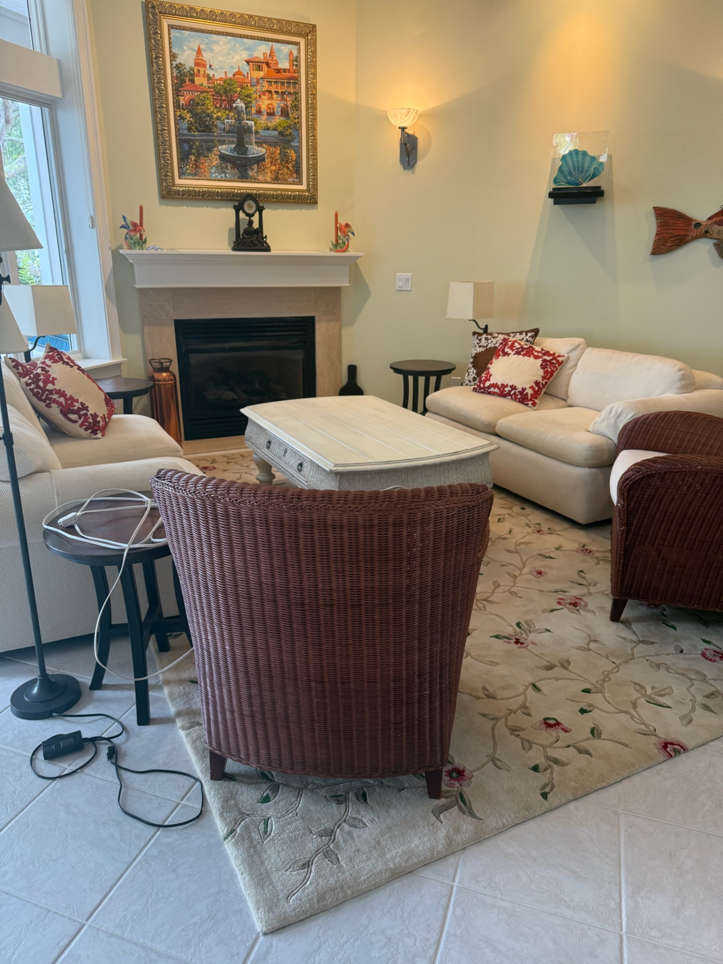 Living room with cream sofas, wicker armchairs, coral pillows, and a fireplace framed by a gilt St. Augustine painting