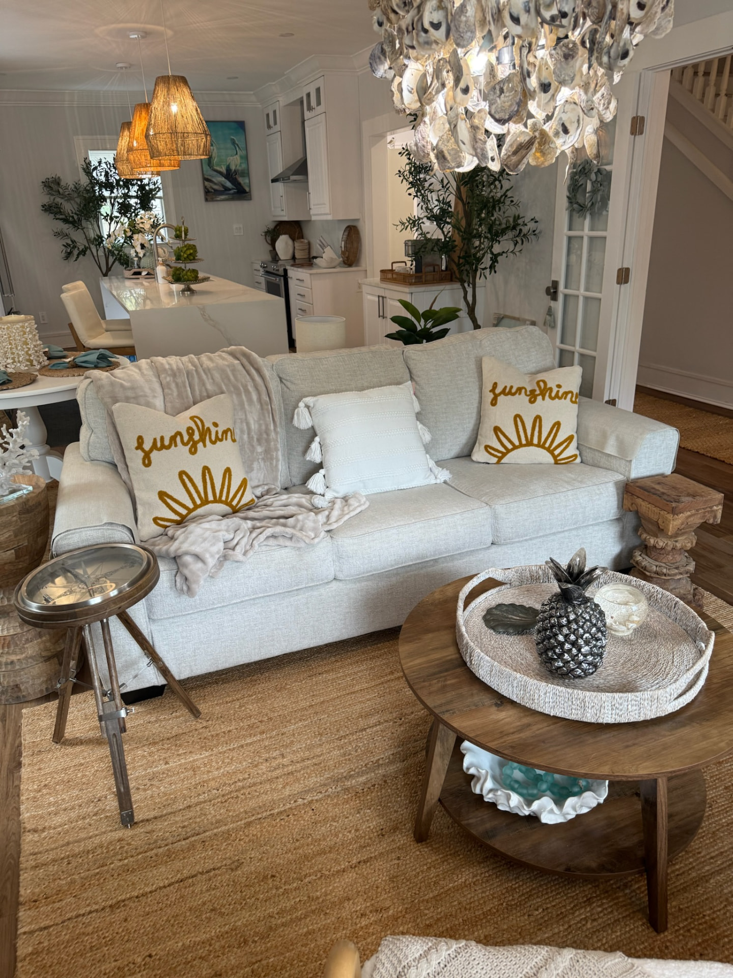 Bright coastal living room with a light gray sofa, sunshine pillows, oyster chandelier, and woven textures