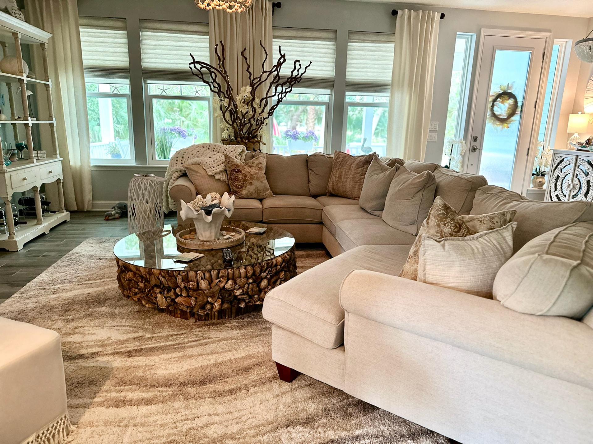 Sunlit coastal living room with a plush sectional, driftwood coffee table, and serene waterfront views by Madeline’s Chic Interiors