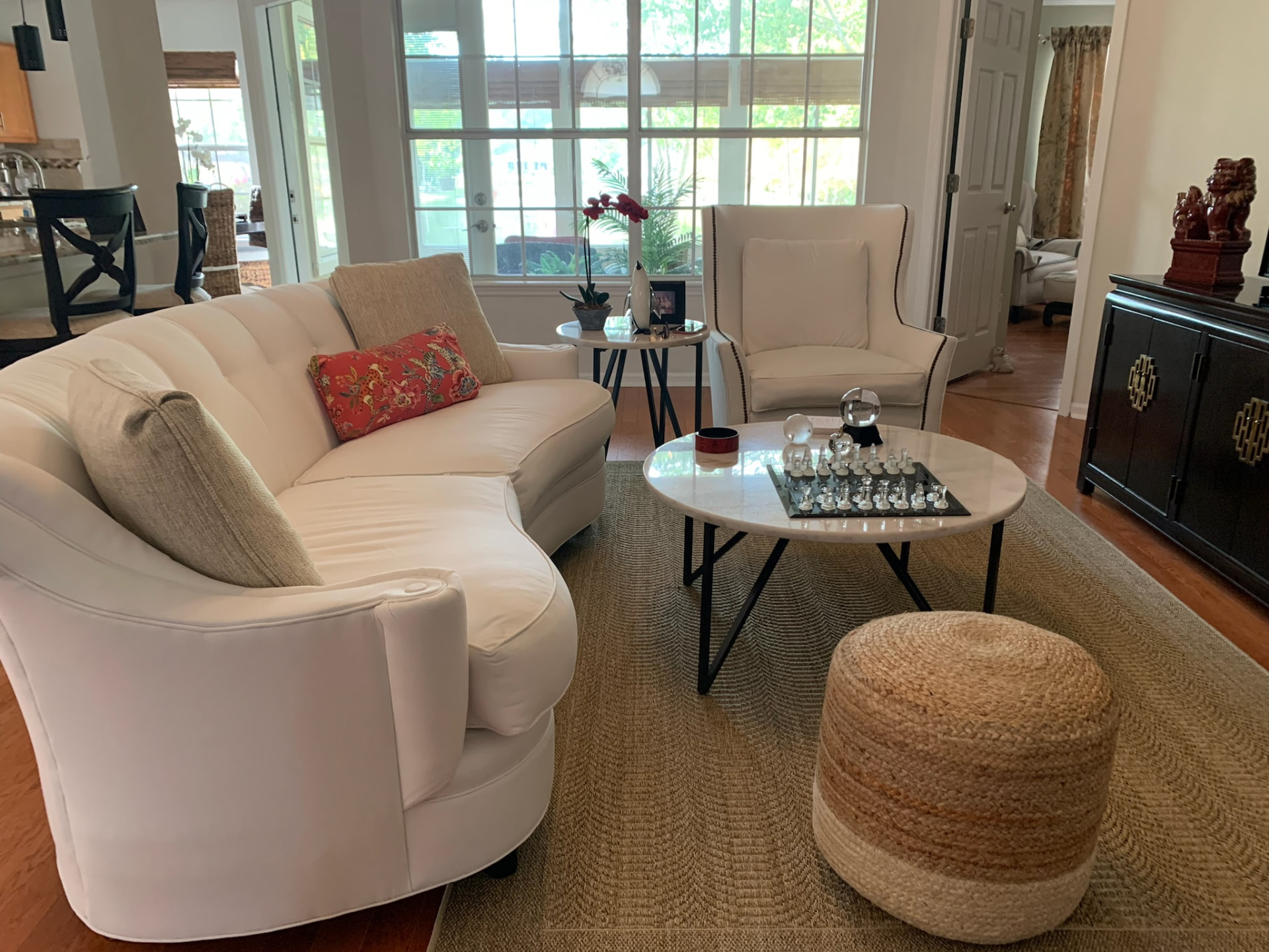 Curved cream sectional with a coral floral pillow, a marble-top coffee table with a chess set, and a woven ottoman in a sunlit living room