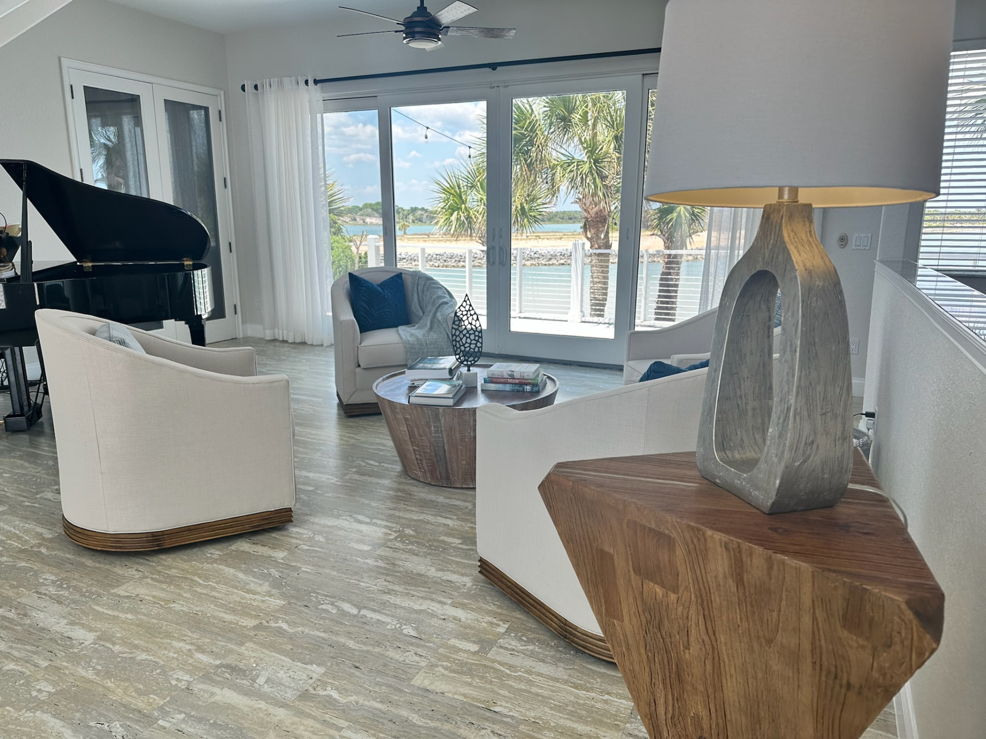 Bright coastal living room with cream sofas, a black piano, and sliding glass doors opening to palm trees and water views
