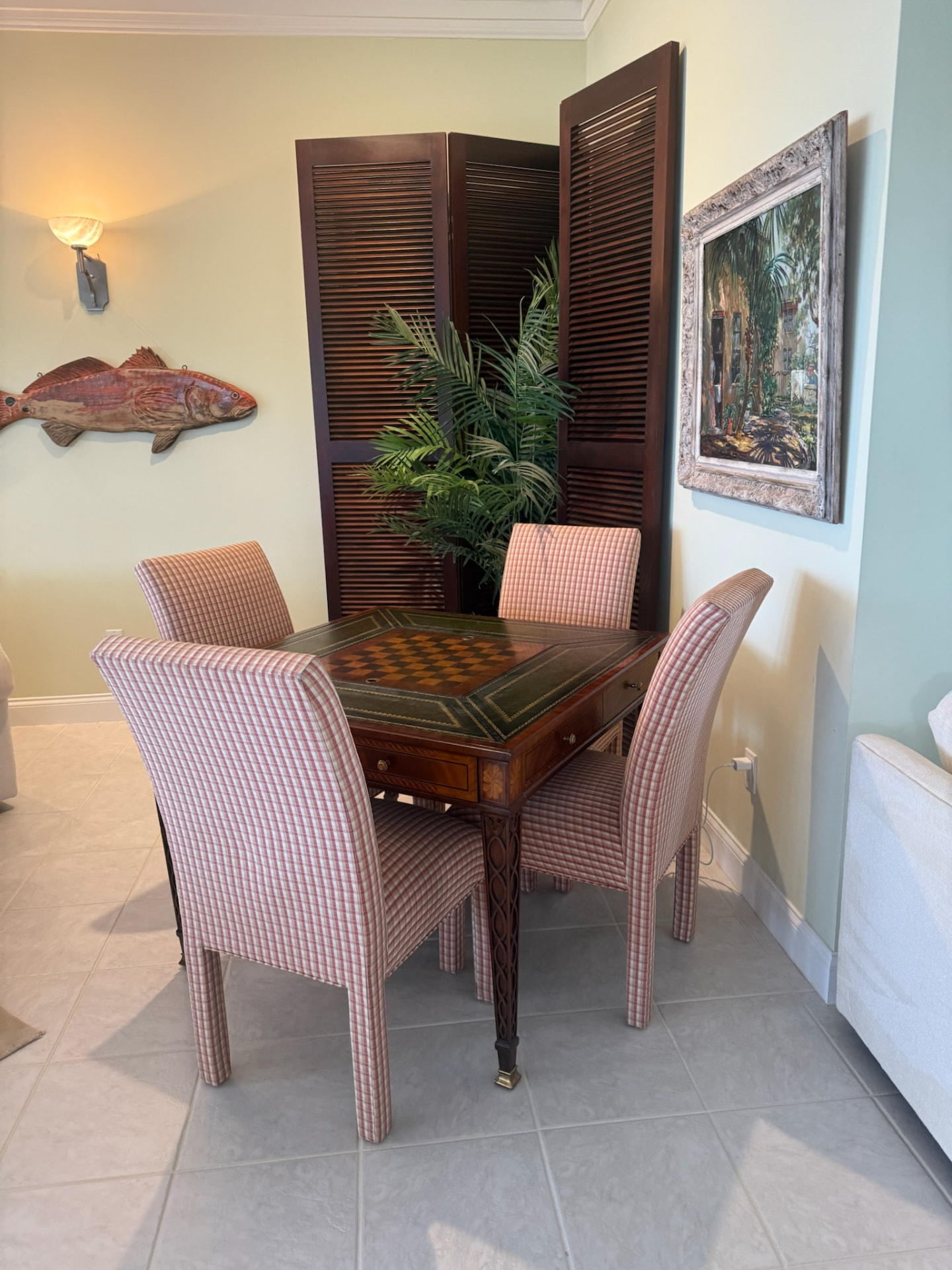 Antique game table with checkerboard inlay surrounded by pink gingham chairs beside a louvered screen and coastal artwork